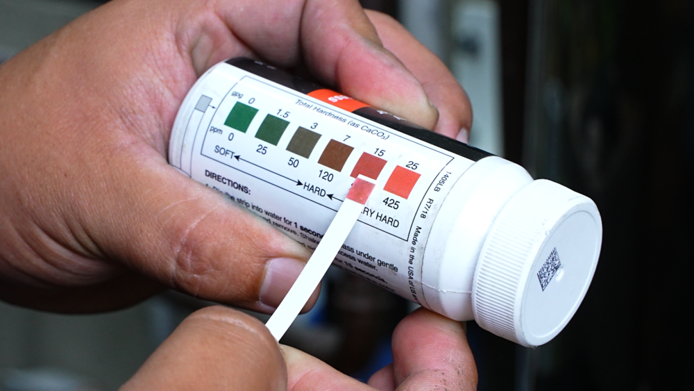 The image shows a technician holding a hard water test strip to the bottle showing hardness scale.