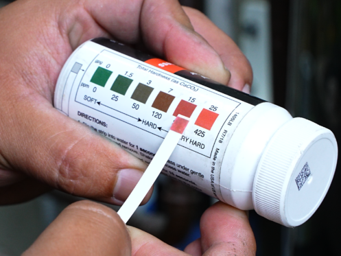 The image shows a technician holding a hard water test strip to the bottle showing hardness scale.