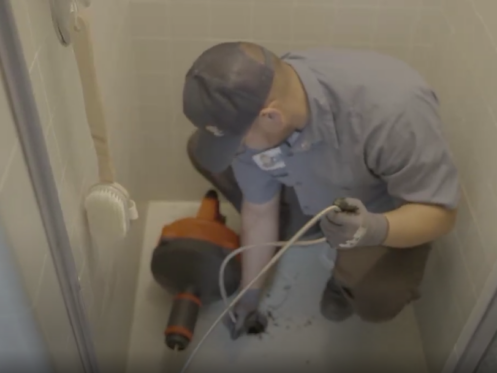 The image shows a Monkey Wrench technician clearing a shower drain with a drain snake.