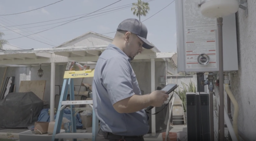 The image shows a Monkey Wrench technician inspecting his work tablet as he performs maintenance on a tankless water heater.