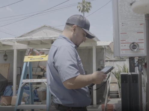 The image shows a Monkey Wrench technician inspecting his work tablet as he performs maintenance on a tankless water heater.