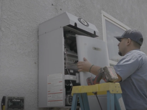 The image shows a Monkey Wrench technician taking the cover off a Noritz tankless water heater.