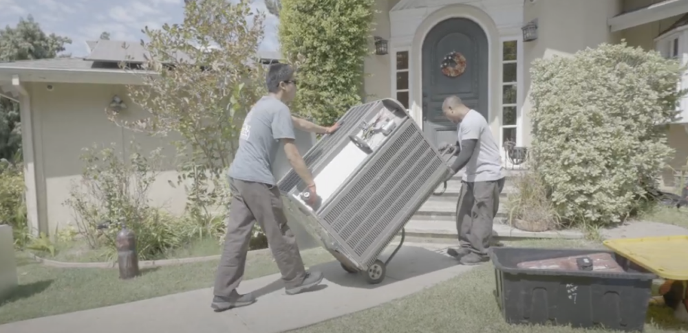 The image shows two Monkey Wrench technicians rolling a HVAC unit into a home.