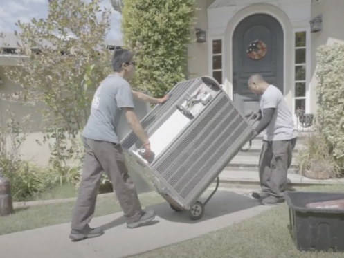 The image shows two Monkey Wrench technicians rolling a HVAC unit into a home.